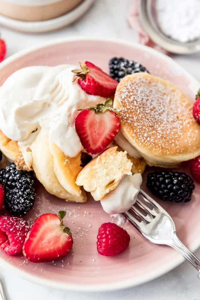 a bite of fluffy Japanese pancake on a fork with whipped cream and fresh berries around it