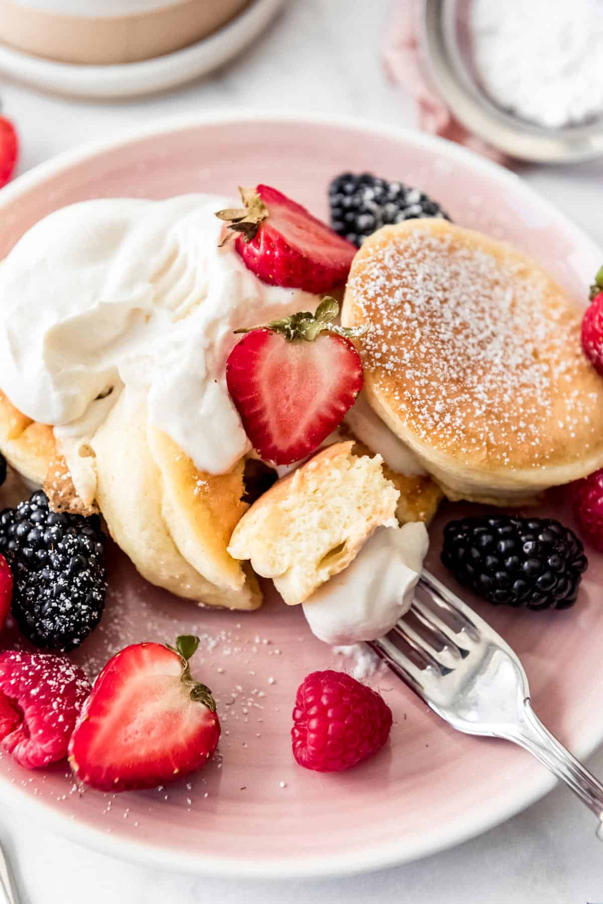 a bite of fluffy Japanese pancake on a fork with whipped cream and fresh berries around it