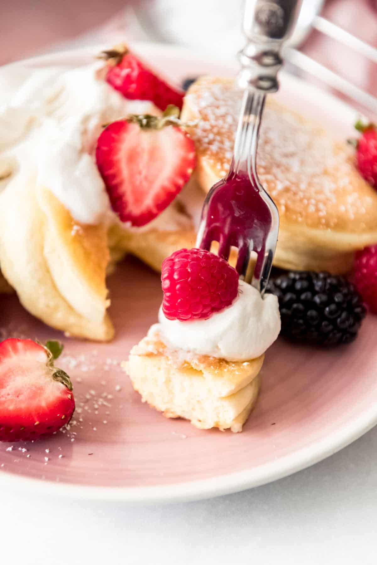 Close up of a bite of a Japanese pancake on a fork, with a dollop of whipped cream and a raspberry. A stack of pancakes is visible in the background, scattered with other berries. 