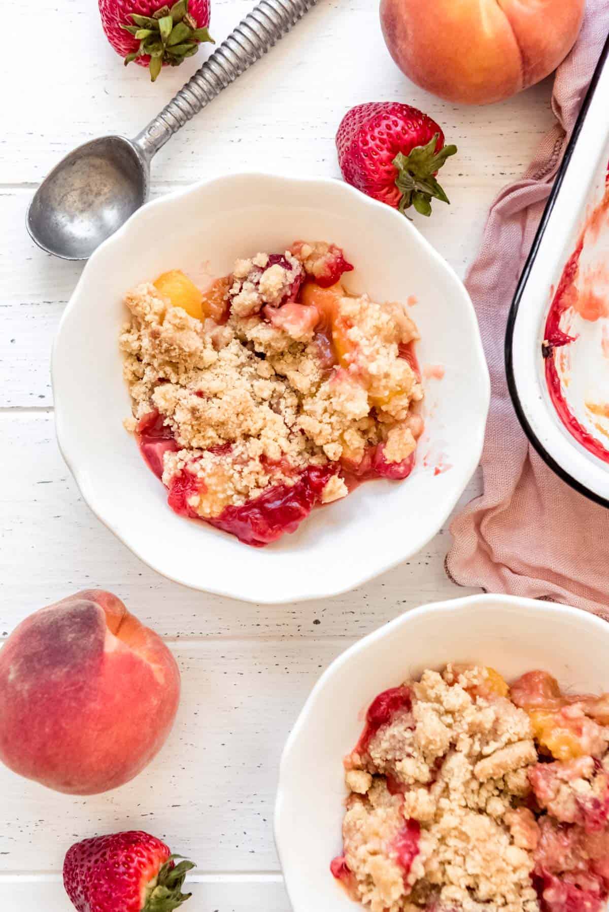 a white bowl of strawberry peach crumble surrounded by fresh strawberries and peaches