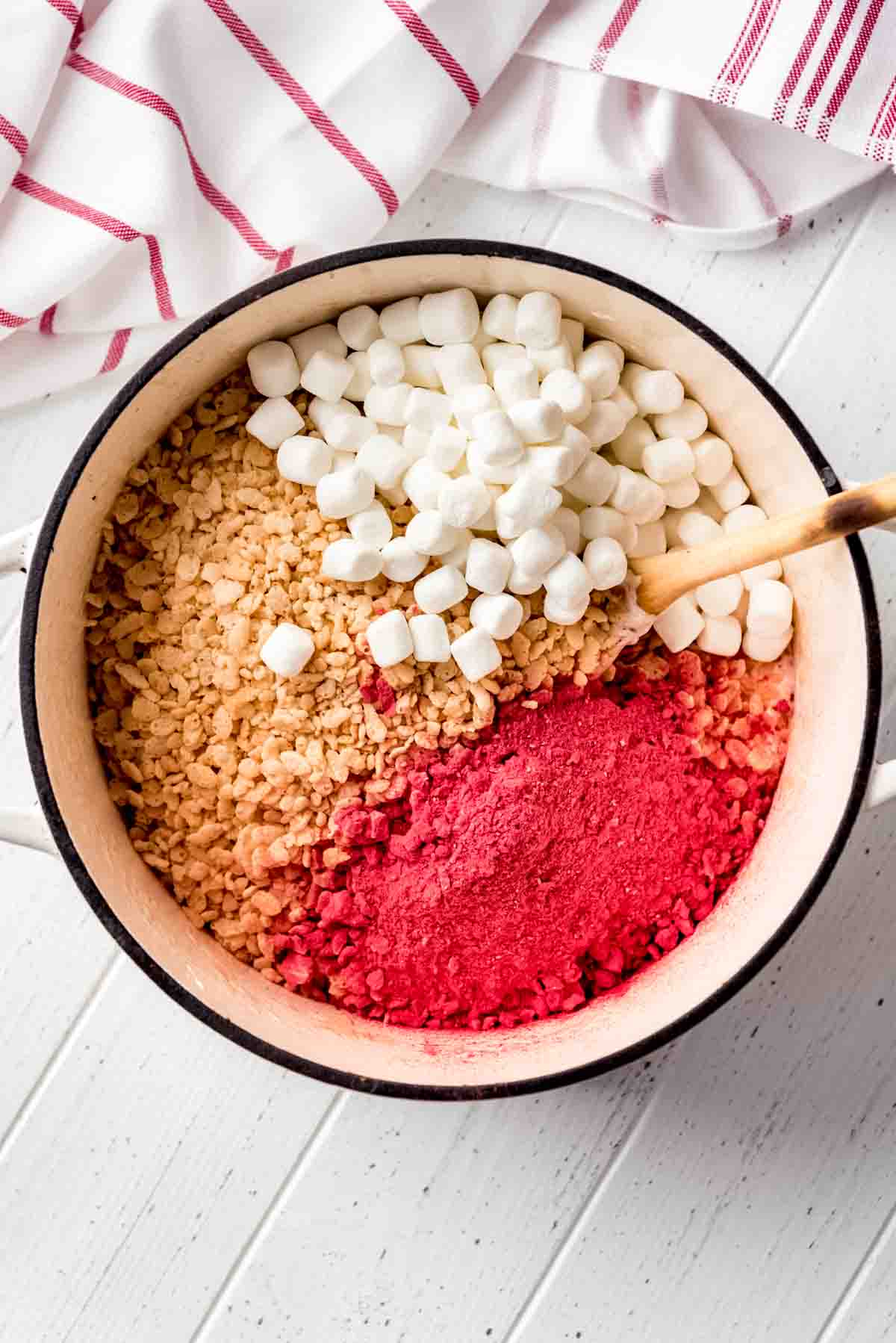 Small marshmallows, rice krispie cereal and crushed strawberry power are in a large pot, ready to be mixed with a wooden spoon that is sticking out of the pot. 