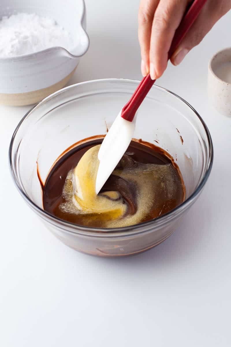 Melted butter and chocolate are stirred with a rubber spatula in a clear glass bowl. 