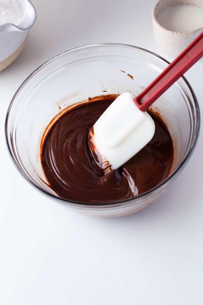 A rubber spatula is resting in a smooth chocolate mixture, in a clear glass bowl. 