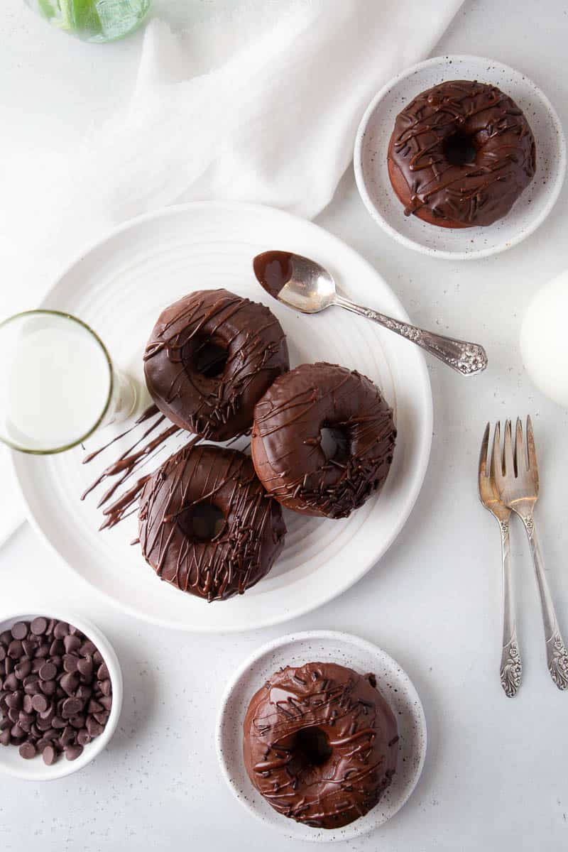 baked chocolate donuts on white plates, a glass of milk, a bowl of chocolate chips, a silver spoon and silver forks