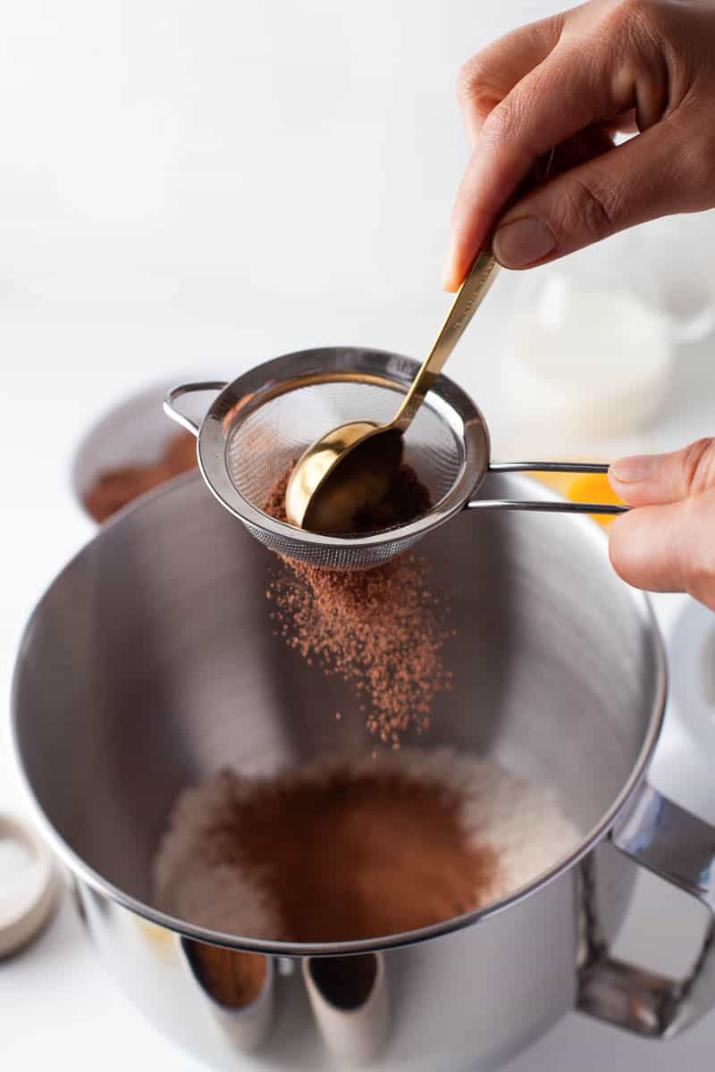 Adding cocoa powder and using a measuring spoon to help sift it into a large bowl. 