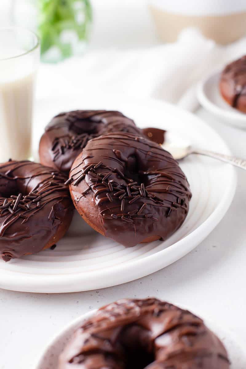 Three glazed chocolate donuts are propped on each other on a white plate with a glass of milk. Other donuts on plates are visible in the foreground and background. 