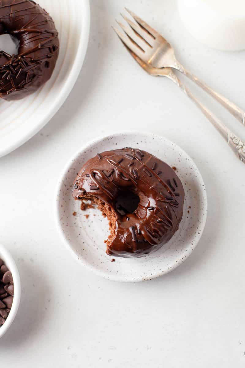 A glazed chocolate donut with a bite out of it is placed on a small plate. Two forks and various bowls are visible around the plate. 