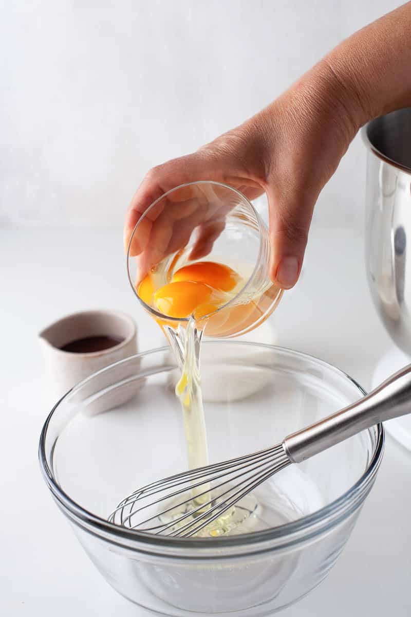 Eggs are added to a clear glass bowl. A wire whisk is resting in the bowl, and other cups and bowls are visible in the background. 