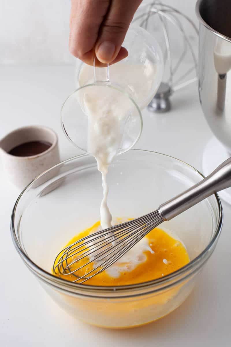 Mike is poured from a small glass pitcher into a clear glass mixing bowl containing eggs. 