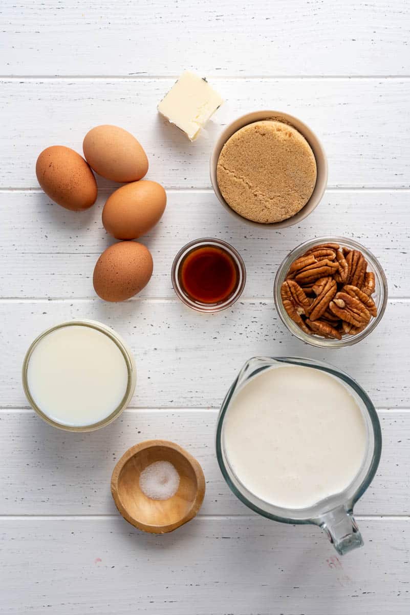 milk, cream, eggs, vanilla, salt, butter, brown sugar, and pecans in separate containers on a white surface