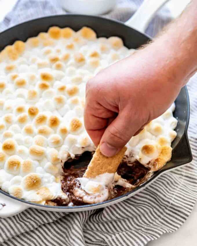 A hand holding a graham cracker stick that's been dipped in melted chocolate and marshmallow displays the treat while a pan with remaining s'mores dip is visible in the background.