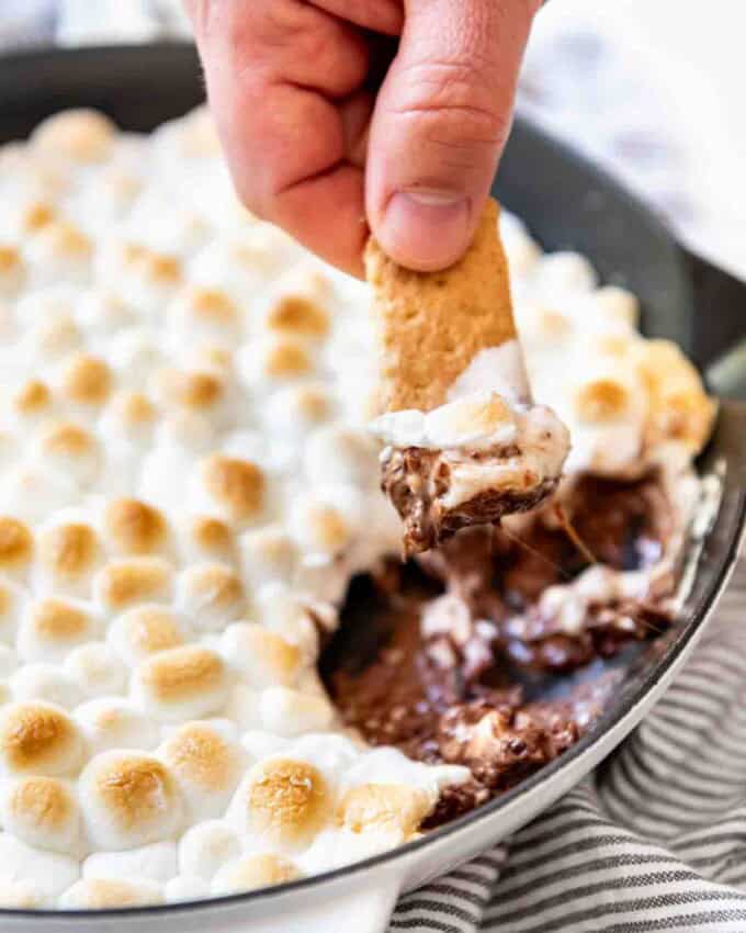 A hand holding a graham cracker stick that's been dipped in melted chocolate and marshmallow displays the treat while a pan with remaining s'mores dip is visible in the background.