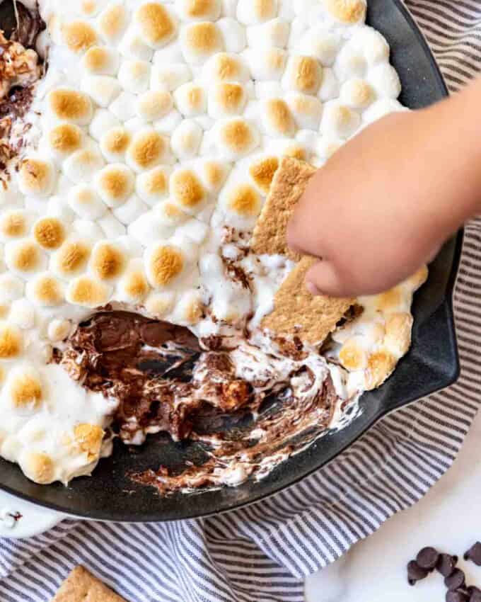 A hand holding a graham cracker to scoop chocolate and marshmallows out of a skillet.