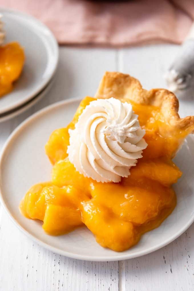 a swirl of sweetened whipped cream on top of a slice of fresh peach pie on a white plate