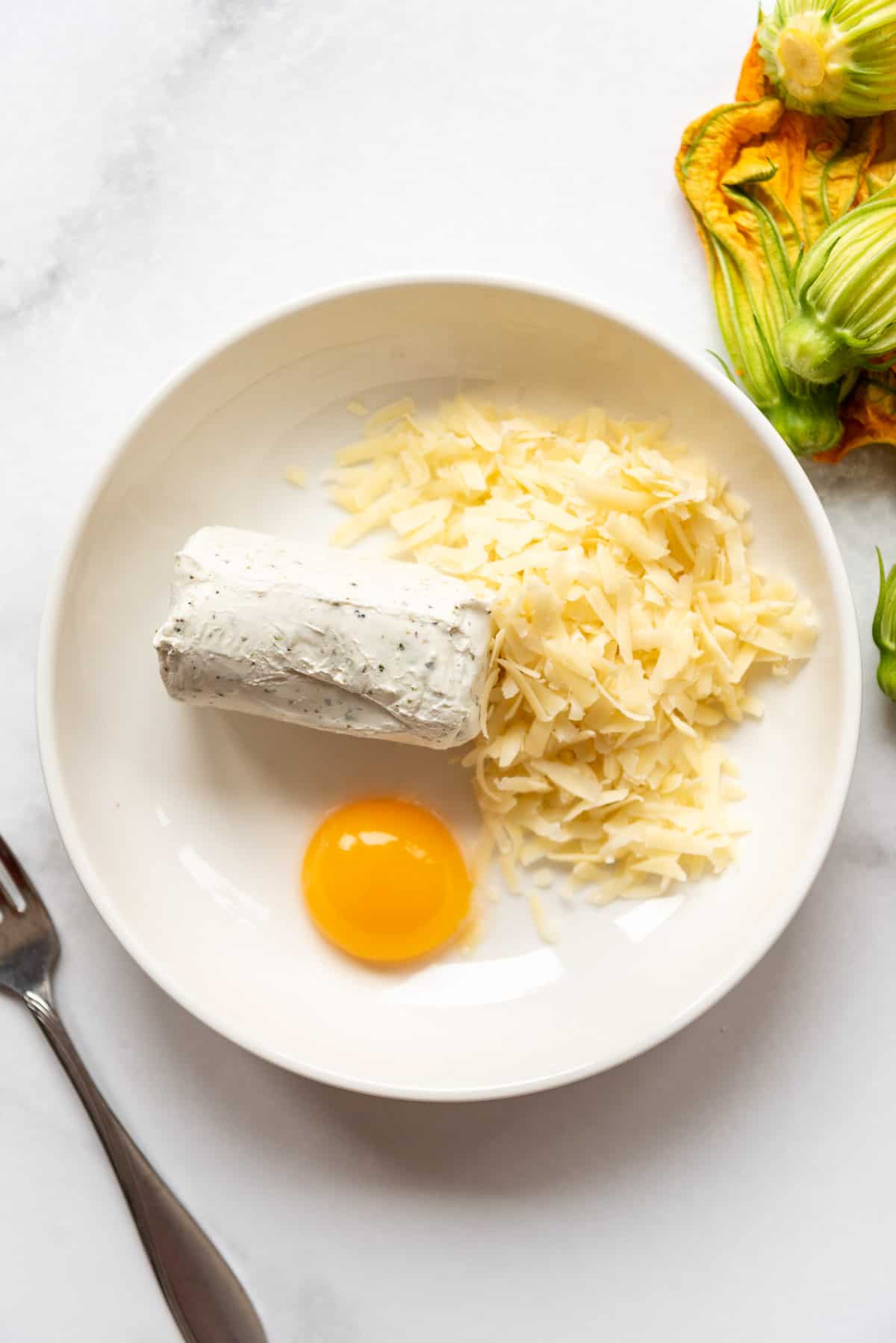 Garlic and herb goat cheese, shredded gruyere cheese, and an egg yolk in a white bowl.