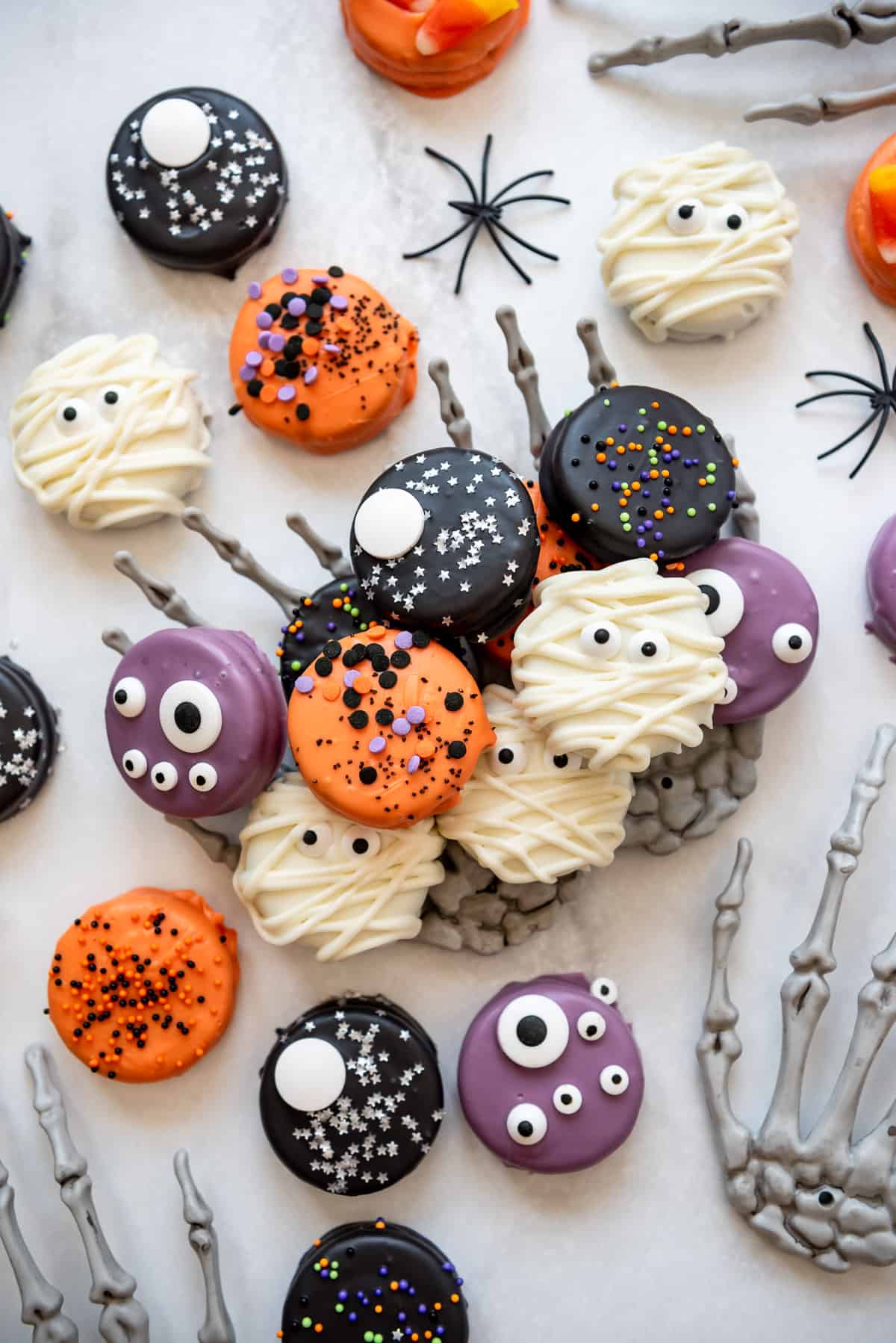 Chocolate covered Oreos decorated in Halloween colors and sprinkles surrounded by plastic spiders and plastic skeleton hands.