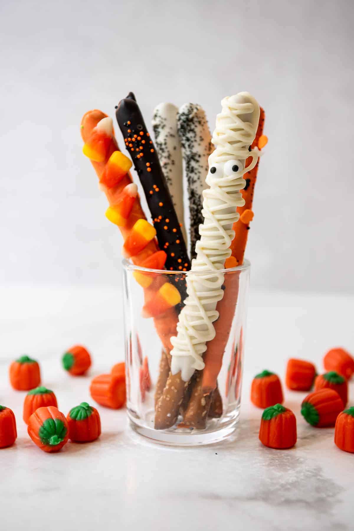 Chocolate covered Halloween pretzel rods displayed in a glass with candy pumpkins next to them.