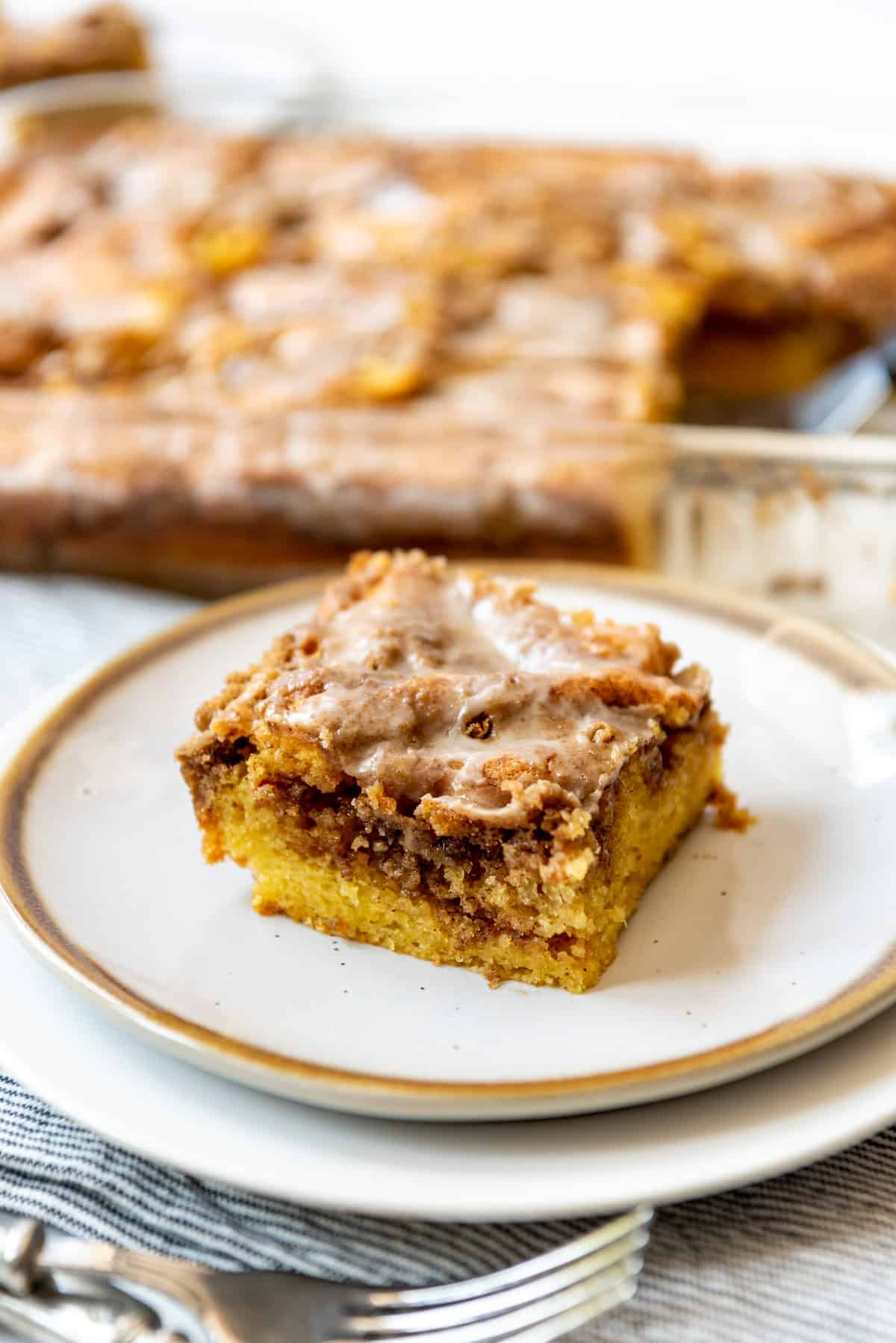 A single slice of cinnamon swirl honey bun cake is on a plate, while a pan containing more cake is visible in the background. 