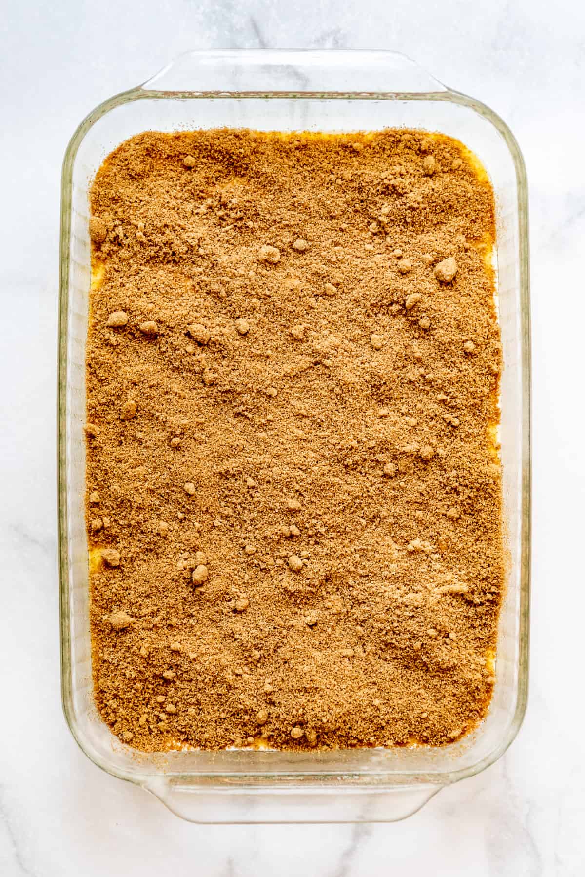 Cinnamon and brown sugar topping is sprinkled on top of honey bun cake batter in a rectangular glass pan. 