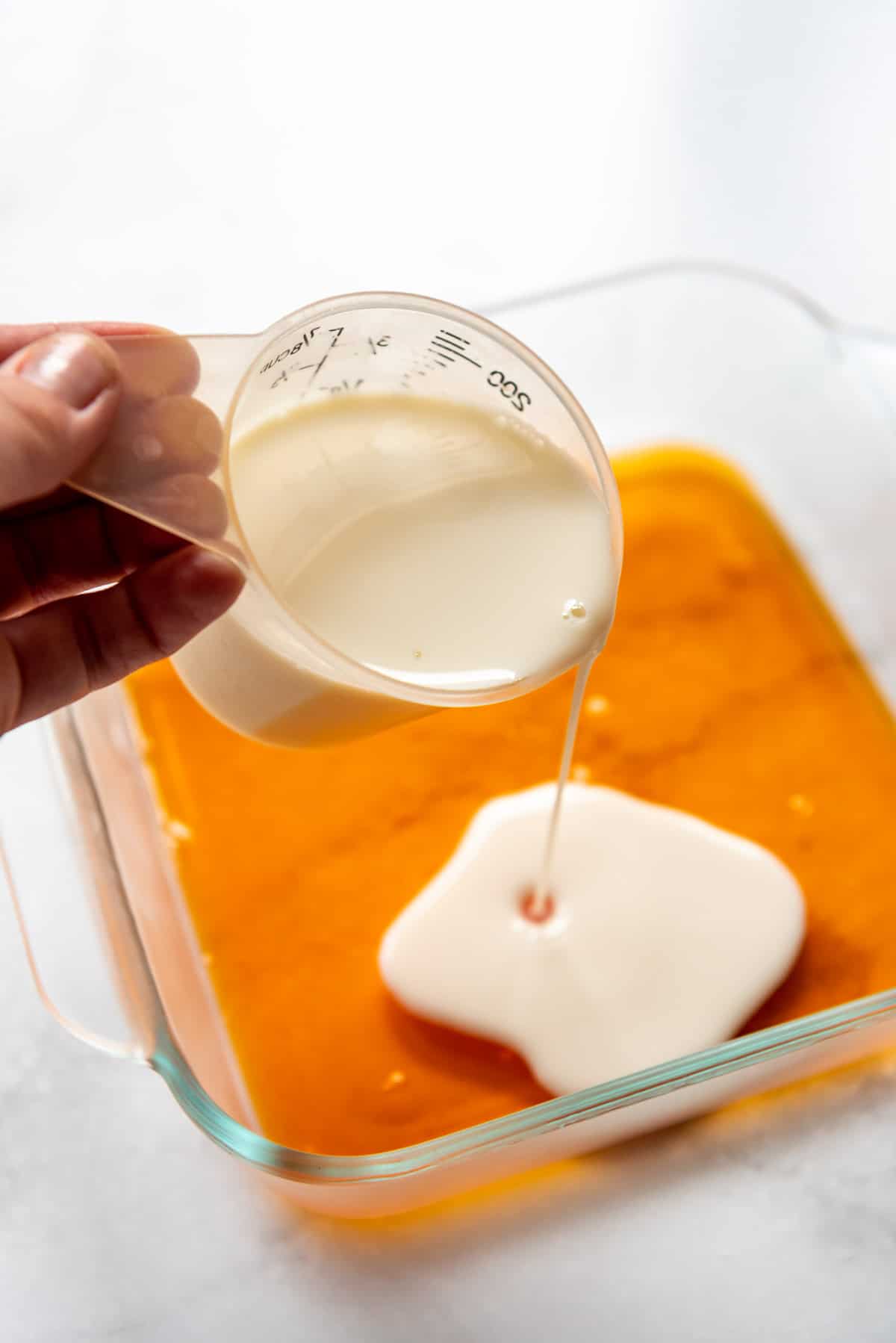 Pouring a layer of white jello liquid onto a set layer of orange jello in a glass dish.