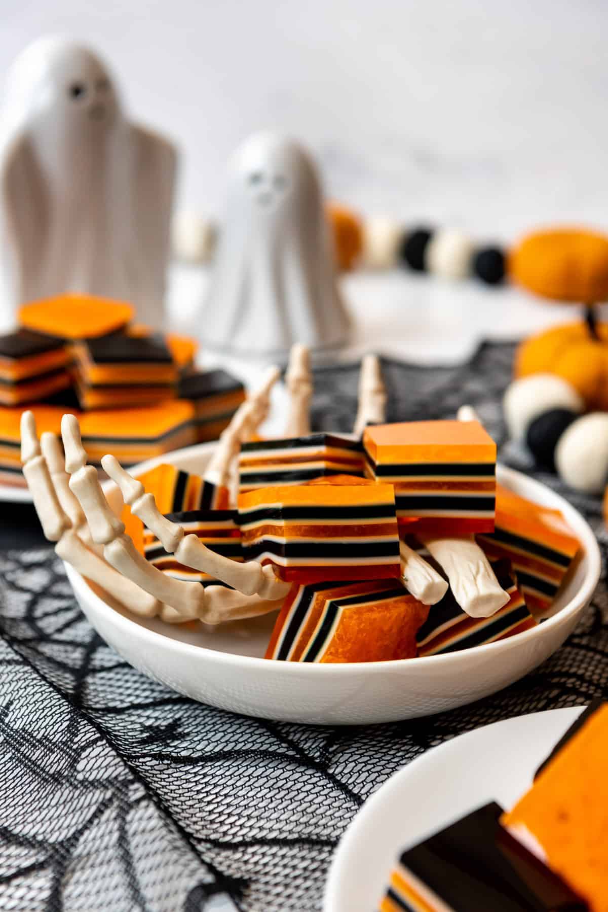 A white dish with plastic skeleton hands holding squares of striped orange, black, and white Halloween jello.