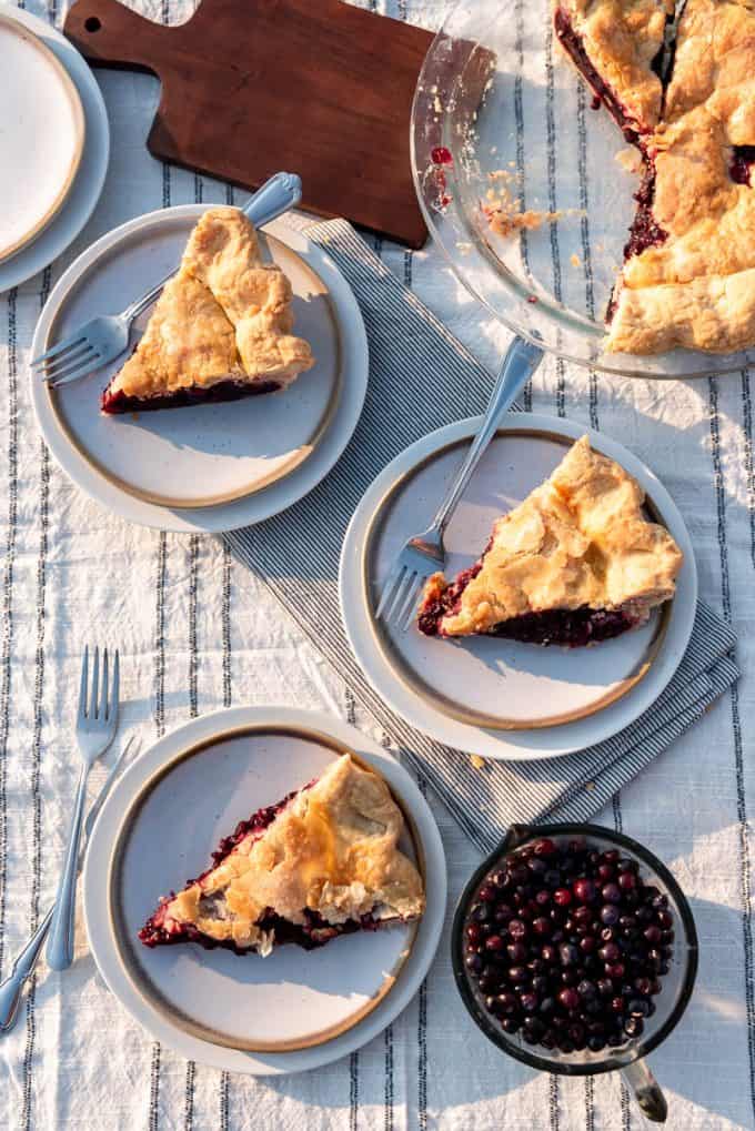 three slices of huckleberry pie on plates next to a glass cup of fresh huckleberries