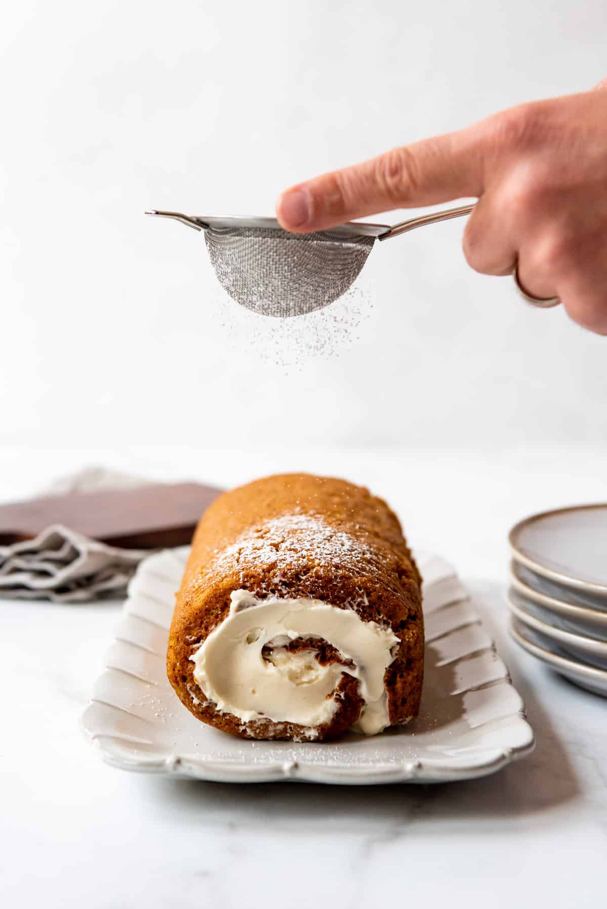 A hand is holding a fine mesh sifter and is dusting the pumpkin roll with powdered sugar. 