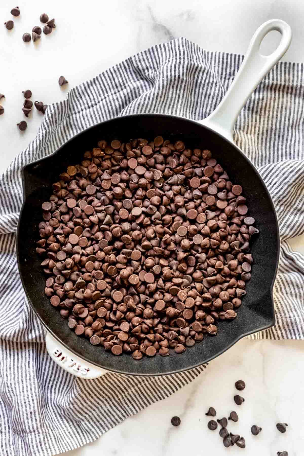 A cast iron skillet holds chocolate chips and is resting on a striped dish towel, with chocolate chips scattered artfully around the sides. 