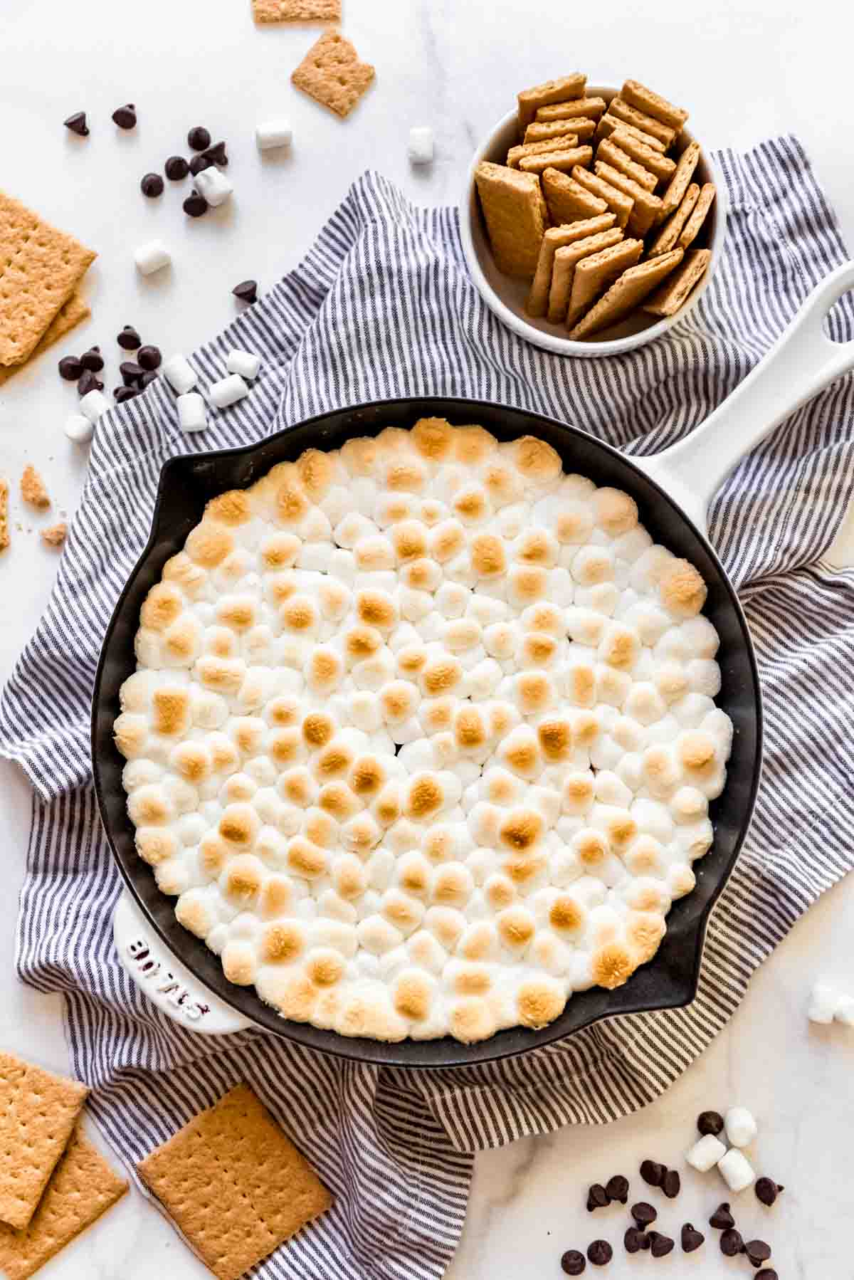 A skillet filled with chocolate and toasted marshmallows next to graham cracker sticks.