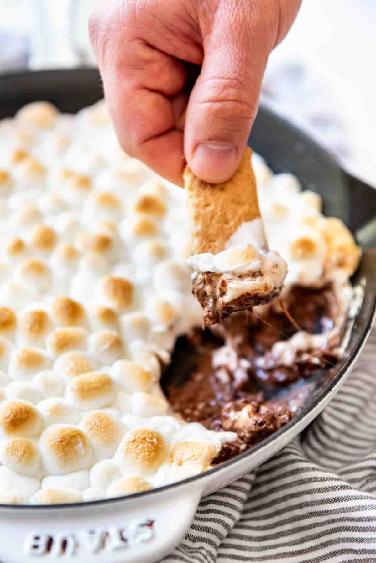 A hand holding a graham cracker stick that's been dipped in melted chocolate and marshmallow displays the treat while a pan with remaining s'mores dip is visible in the background. 