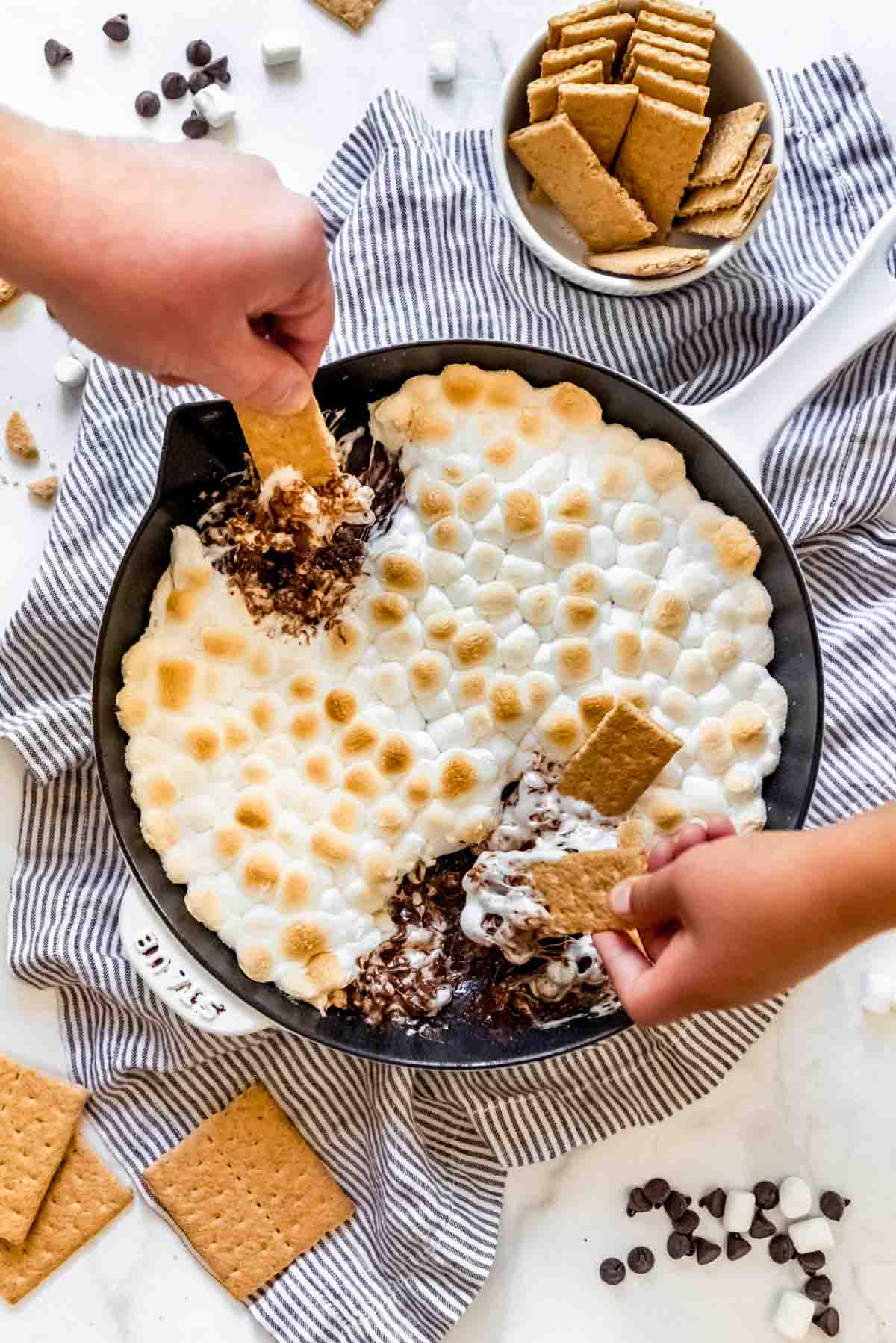 Hands scooping melted chocolate and toasted marshmallows out of a skillet with graham crackers.