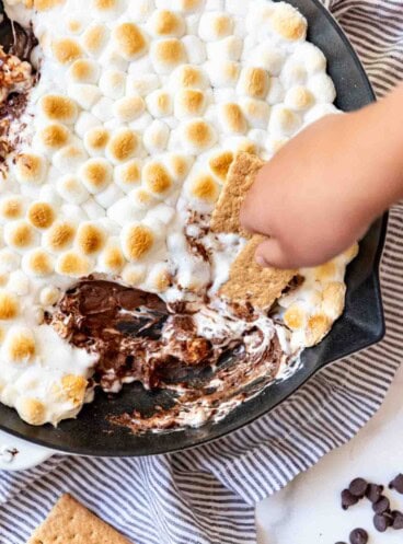 A hand holding a graham cracker to scoop chocolate and marshmallows out of a skillet.