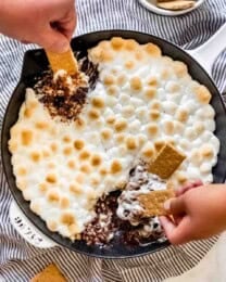 Hands scooping melted chocolate and toasted marshmallows out of a skillet with graham crackers.