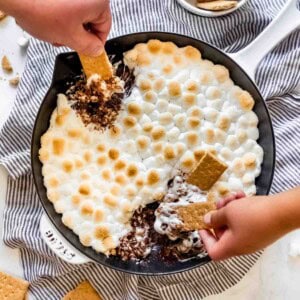 Hands scooping melted chocolate and toasted marshmallows out of a skillet with graham crackers.