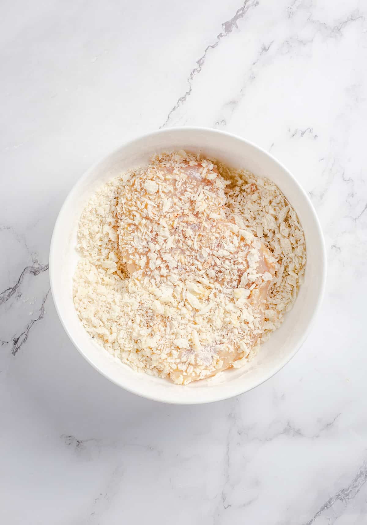 A chicken breast being dredged in Panko breadcrumbs in a white bowl.