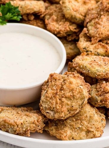 A bowl of ranch dipping sauce next to fried pickles stacked on a white plate.