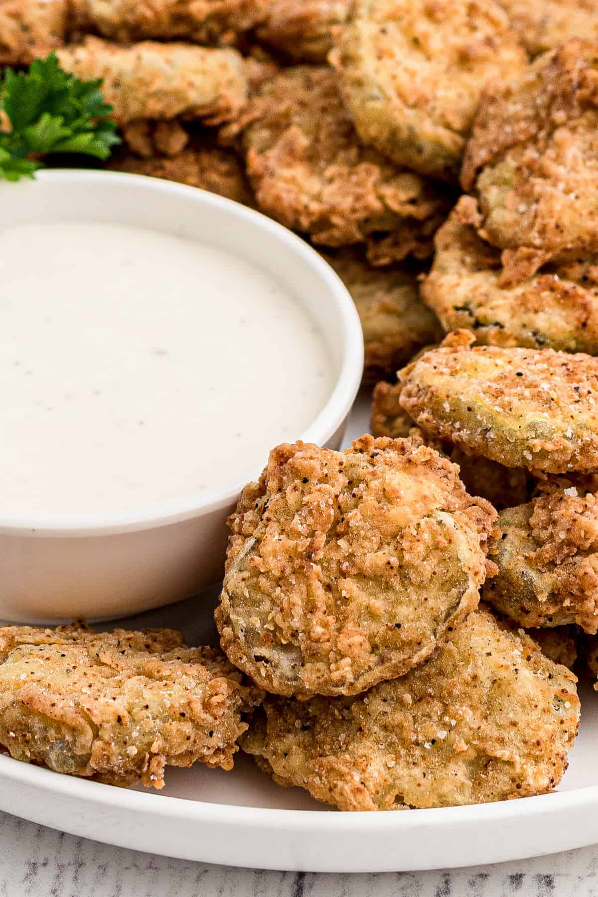A bowl of ranch dipping sauce next to fried pickles stacked on a white plate.