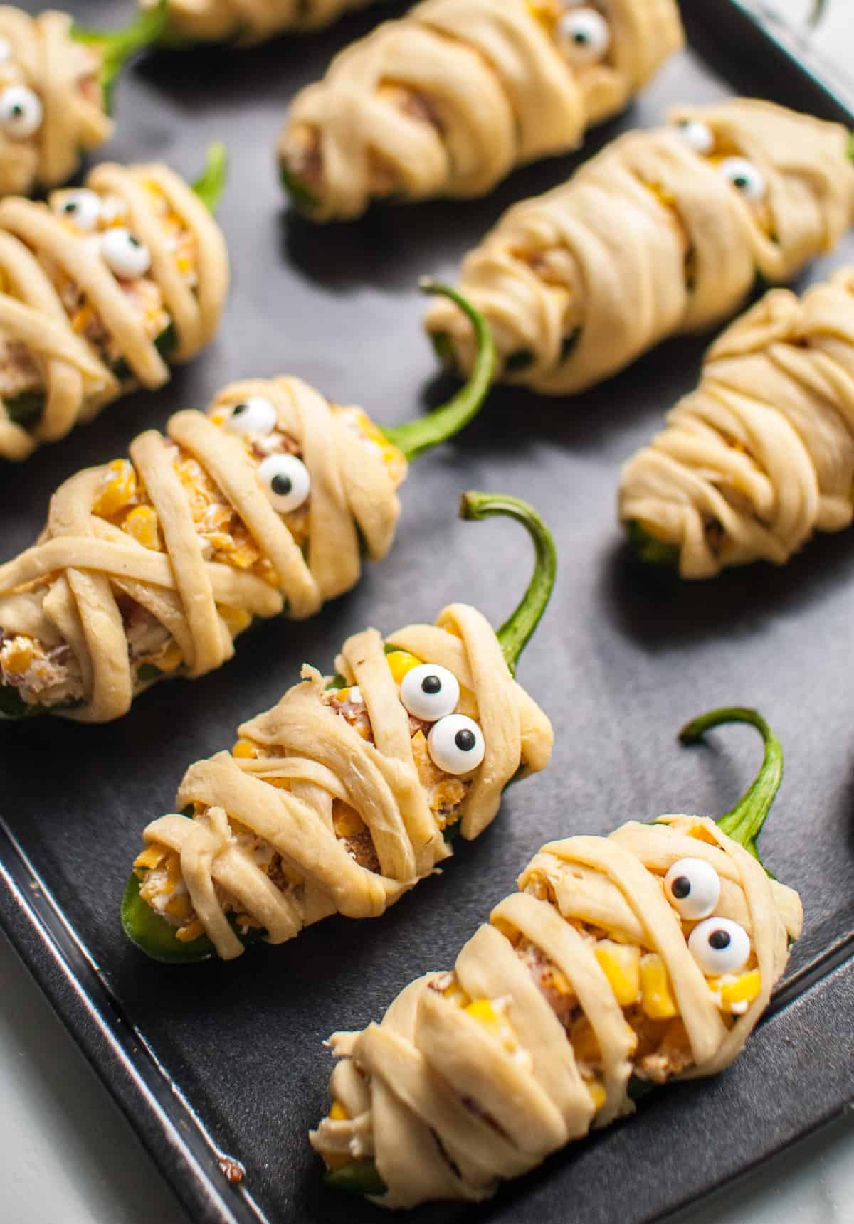 Mummy jalapeno poppers lined up on a baking sheet ready to be baked.