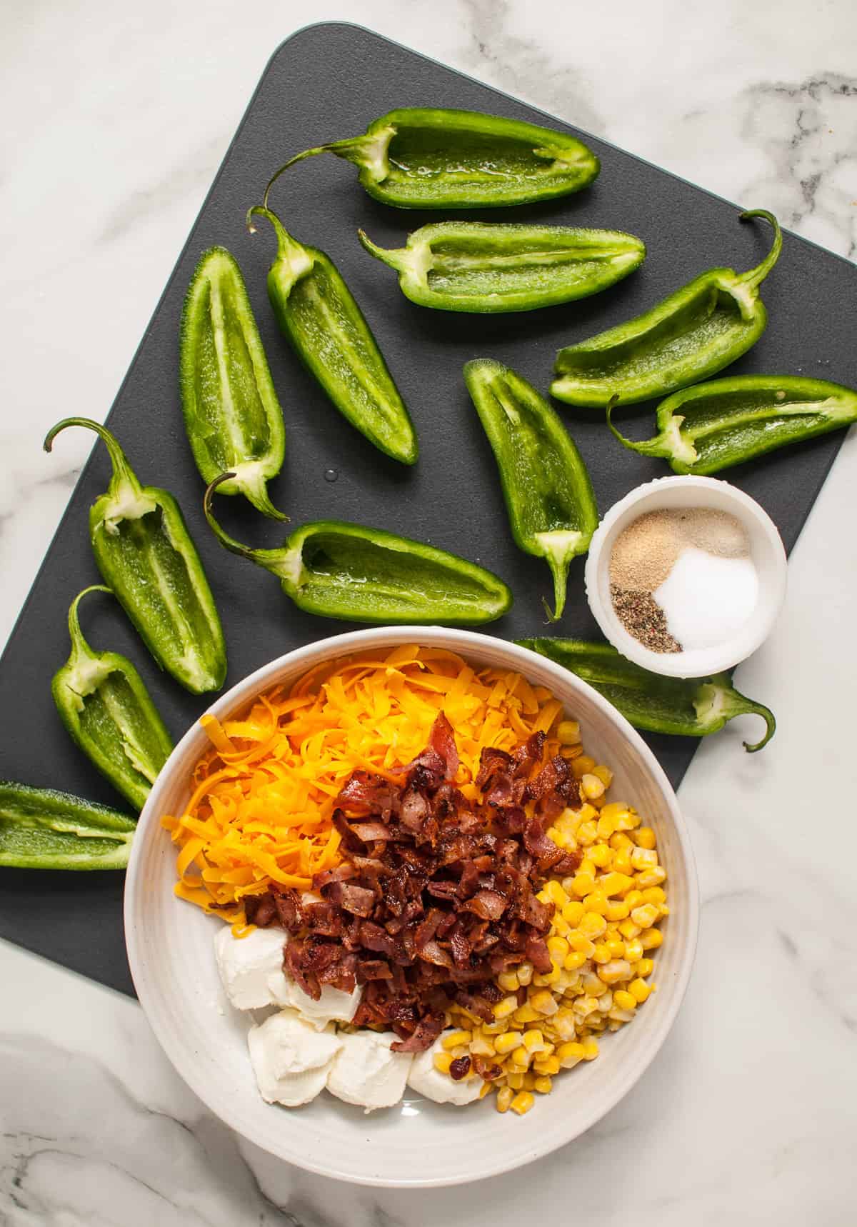 Cream cheese, crumbled bacon, cheddar cheese, and corn in a bowl next to cleaned and sliced jalapeno halves.