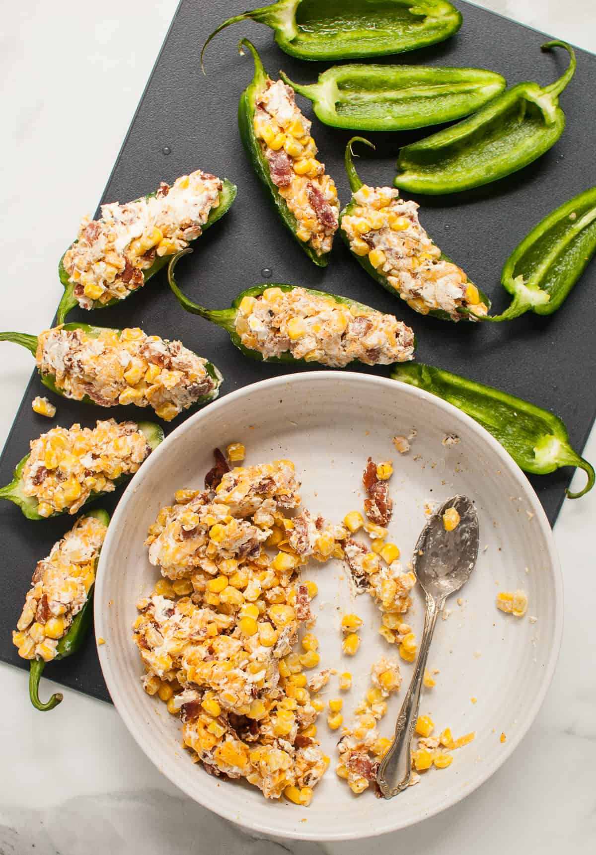 A bowl of corn and cream cheese filling next to jalapeno halves, some of which have been filled.