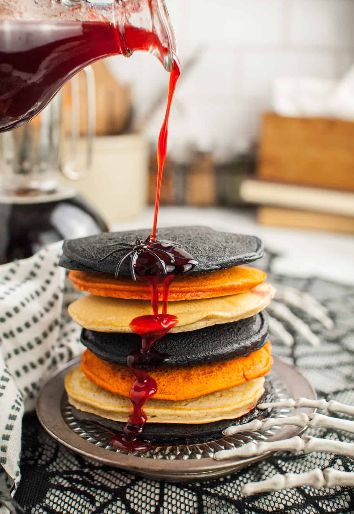 Red strawberry syrup being poured over a stack of black, orange, and white pancakes for a Halloween breakfast.