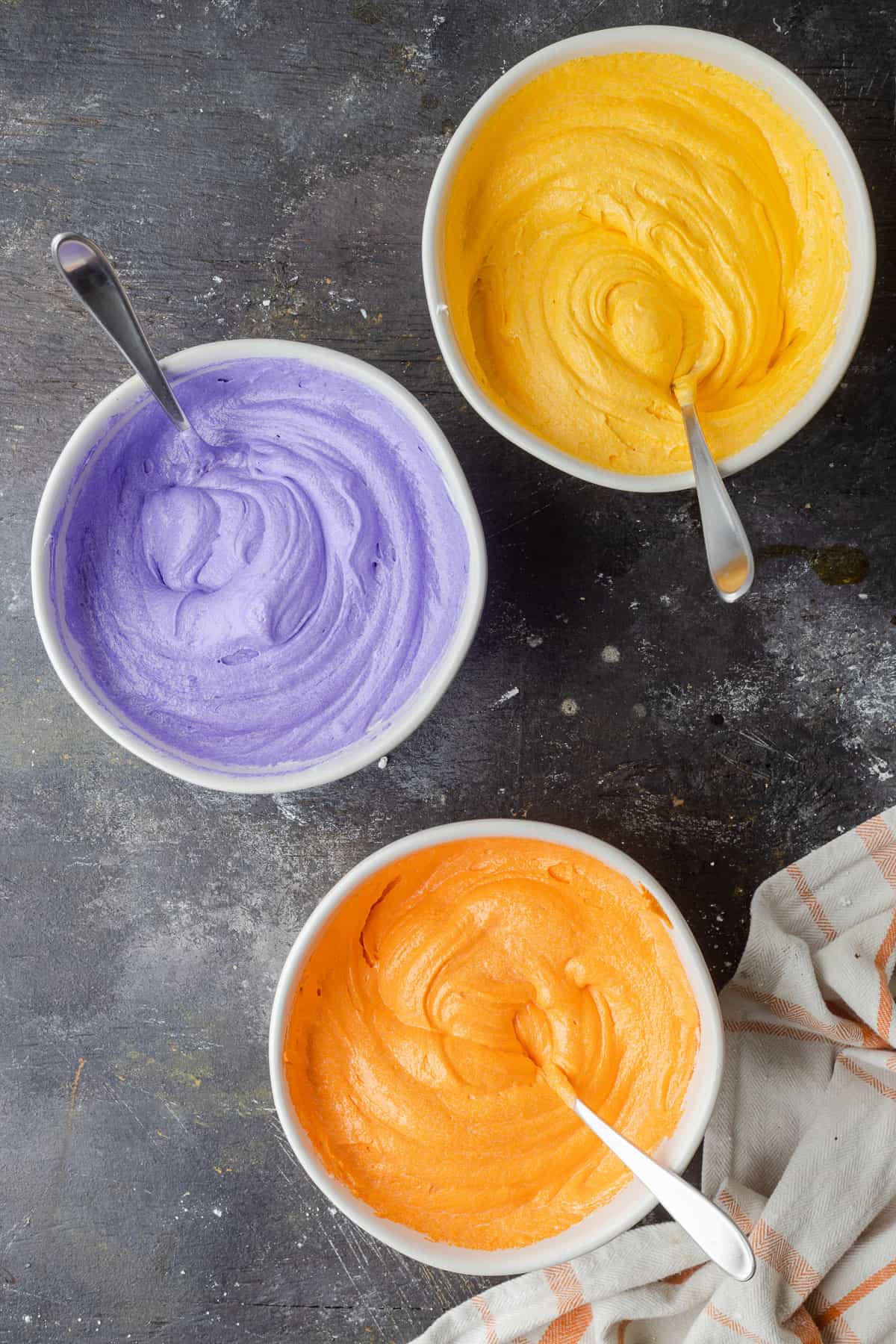 Adding yellow, purple, and orange food coloring to each bowl of frosting to make three different colors.