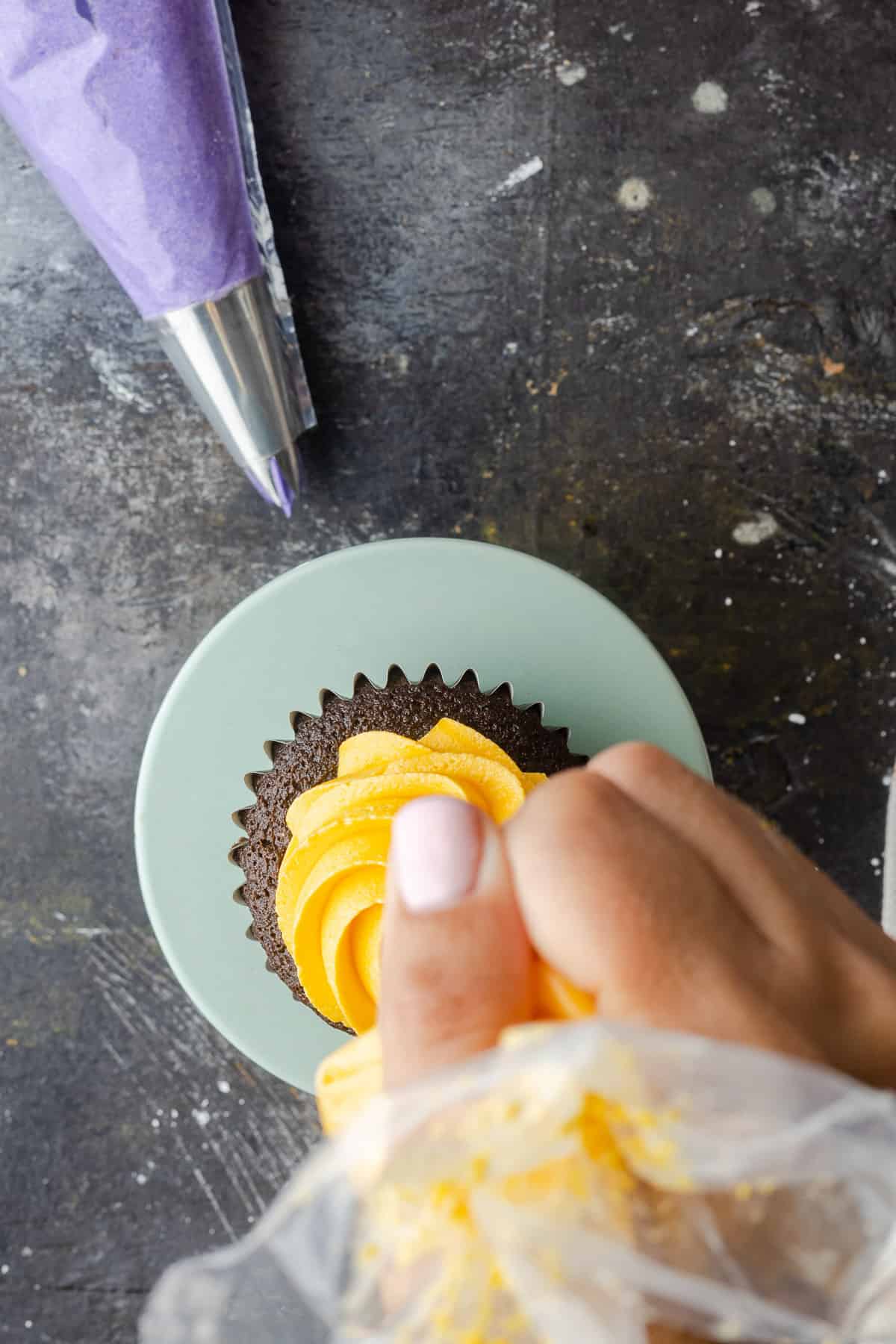 A hand piping a swirl of yellow frosting onto a cupcake.