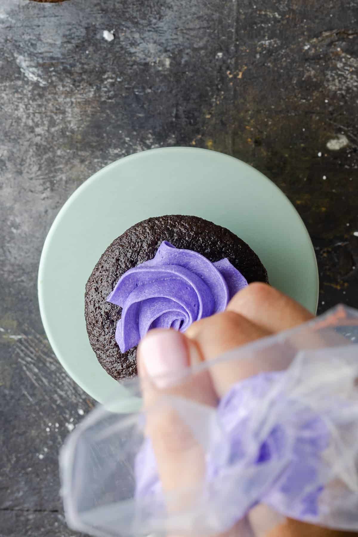 A hand piping a swirl of purple buttercream frosting onto a chocolate cupcake.