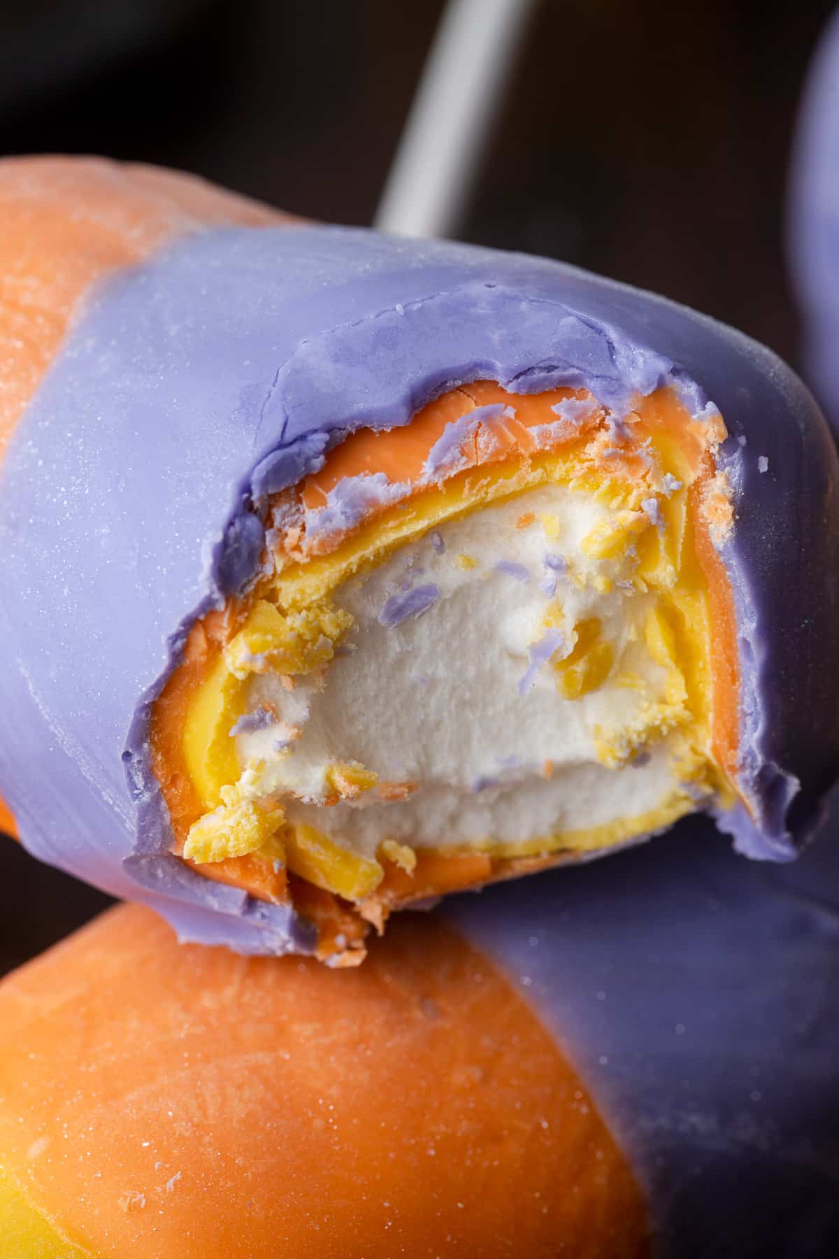 a bite taken out of a marshmallow pop covered in yellow, orange, and purple white chocolate.