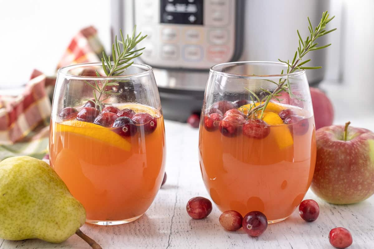 Two glasses of cider decorated with rosemary sprigs, fresh cranberries, and orange wedges.