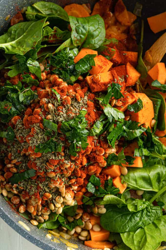 Top view of a black pot with sauteed vegetables and green leaves, lentils, paprika and oregano, with fresh parsley on top.