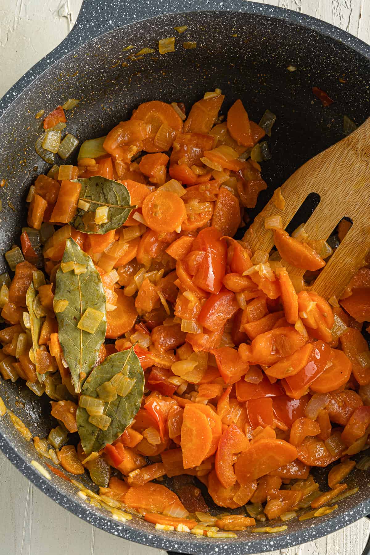 Top view of a black pot with sauteed carrots, onion, garlic, and chopped tomatoes in it with bay leaves mixed in.