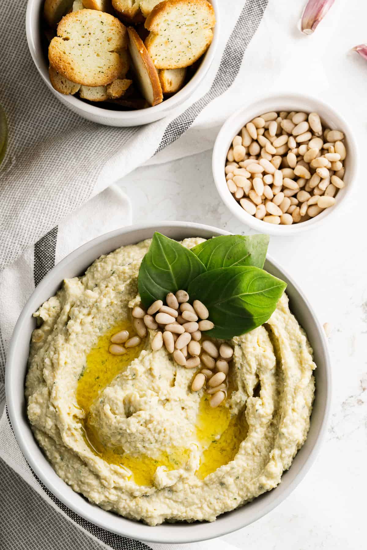 A large bowl of homemade hummus with pesto flavors, pine nuts, and fresh basil leaves on top.