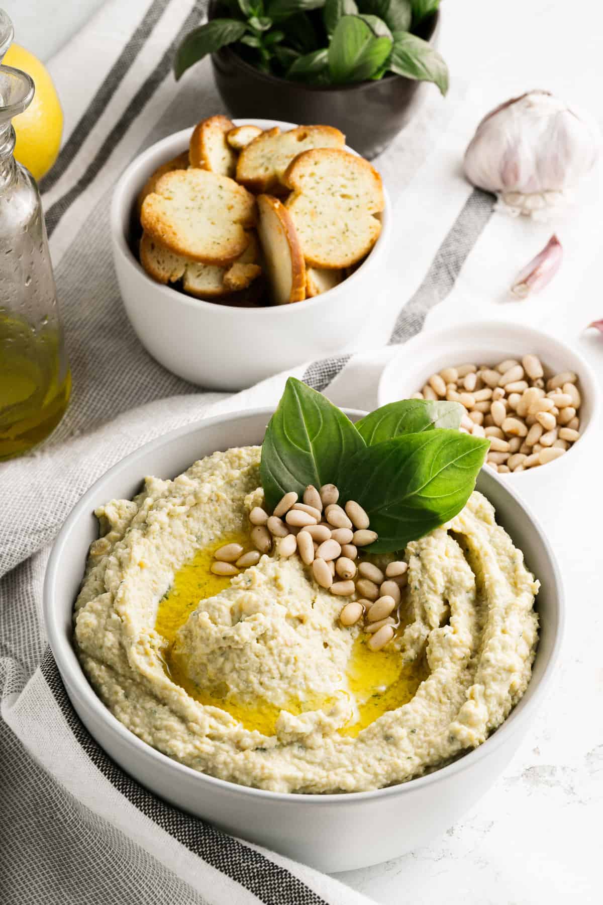 A white bowl filled with pesto hummus in front of a bowl of pine nuts and a bowl of toasted bagel chips.